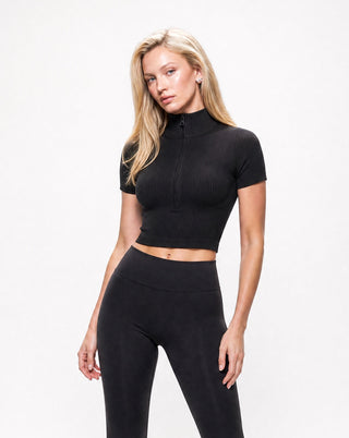 Women's black ribbed zip-front crop top with a mock neckline, short sleeves, and sculpting fit, styled with high-waisted leggings for a sleek athleisure look