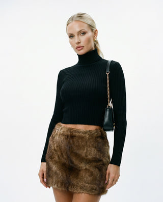 Model wearing a fitted black ribbed long sleeve turtleneck crop top paired with a brown faux fur mini skirt and a black shoulder bag with gold chain strap, photographed against a clean white studio backdrop.