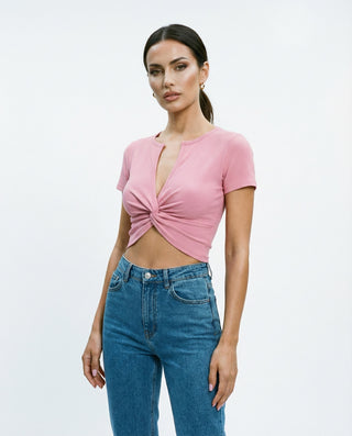 Model wearing a pink ribbed short-sleeve crop top with twisted front detail and deep V neckline, styled with high-waisted blue jeans