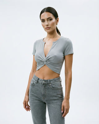 Model wearing a stone gray ribbed short sleeve crop top with a front twist detail and subtle plunge neckline, paired with high waisted gray denim jeans, photographed in a white studio setting.