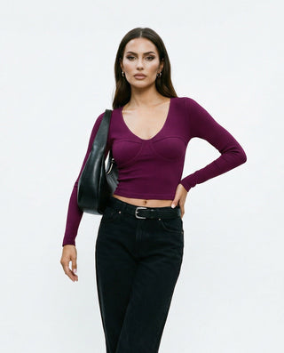 Angled view of a women’s burgundy bustier-style crop top showing contoured seams, long sleeves, and a body-hugging fit paired with black jeans.