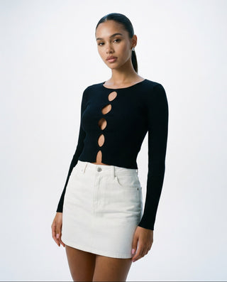 Model styled in a fitted black long sleeve crop top featuring vertical oval cutouts down the center front, paired with a white mini skirt.
