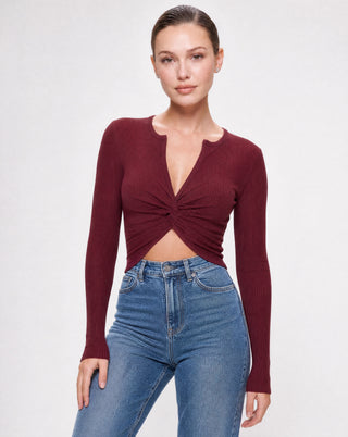 Women's burgundy ribbed long sleeve crop top featuring a twist front detail, V-neckline, front cutout, and fitted silhouette styled with high waisted blue jeans