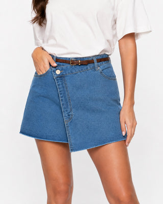Close-up of women’s medium wash denim wrap skort with asymmetrical front, button closure and raw hem, styled with a white T-shirt and brown belt for an effortless casual everyday outfit.