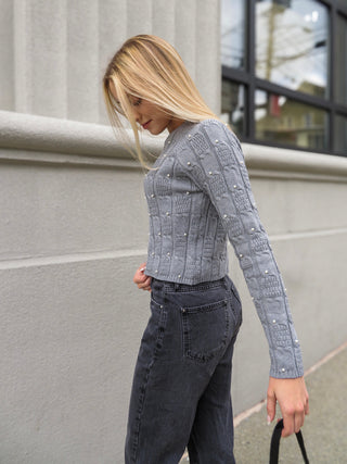 Woman wearing gray quilted sweater adorned with pearls and black jeans. She is hold a black bag.