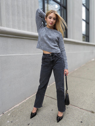 Woman wearing gray quilted sweater adorned with pearls and black jeans. She is holding a black bag and sporting black and gold heels.